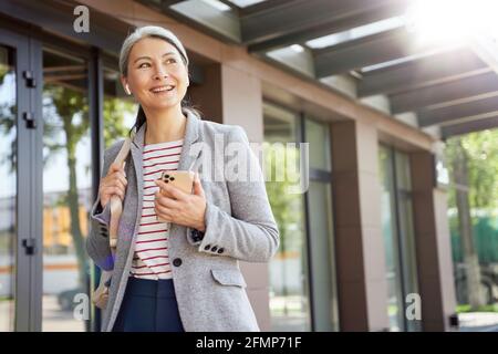Portrait d'une femme belle et heureuse portant des écouteurs sans fil à l'aide d'un smartphone, parlant avec quelqu'un par téléphone en étant debout à l'extérieur Banque D'Images