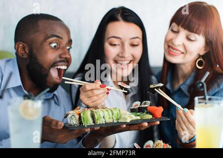 Trois jeunes gens multiethniques, enthousiastes et affamés, mangeant des petits pains à sushis japonais au restaurant avec bonheur et joie. Gros plan. Concept de réunion d'amis et de sushi Time Banque D'Images