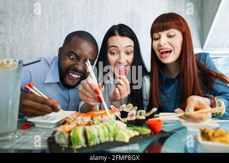 Les gens, la nourriture, le plaisir, le repas de sushi. Gros plan de trois amis multiethniques affamés et excités, s'amuser dans un café, manger de la nourriture asiatique, prendre des petits pains à sushis avec des bâtonnets de bambou Banque D'Images