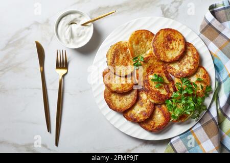 Crêpes de pommes de terre au fromage cottage à l'ail, persil, servies avec une sauce à la crème sure sur une assiette sur fond de pierre en marbre clair avec couverts dorés Banque D'Images