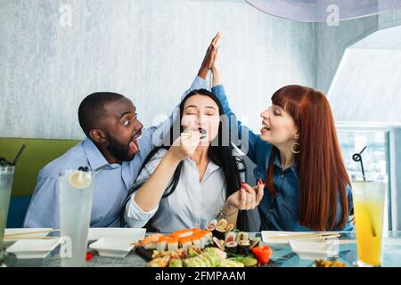 La nourriture savoureuse, le plaisir de manger. Jolie jeune femme asiatique appréciant le délicieux sushi roll, assis à la table au café, tandis que ses amis, l'homme africain et la femme caucasienne donnant élevé cinq l'un à l'autre Banque D'Images