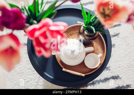 Décoration intérieure et intérieure. Fleurs de tulipes fraîches mises dans des vases sur une table à café avec bouilloire à thé et une tasse sur un plateau en rotin vue du dessus Banque D'Images