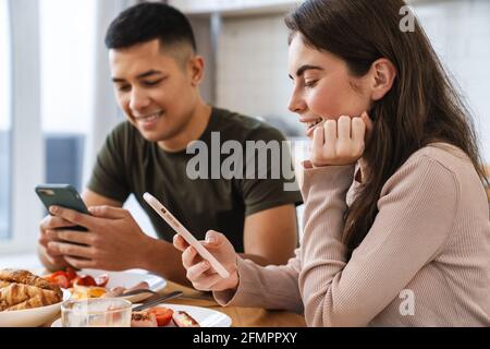 Portrait d'un couple souriant utilisant un smartphone et parlant tout en prenant le petit déjeuner dans une cuisine confortable à la maison Banque D'Images