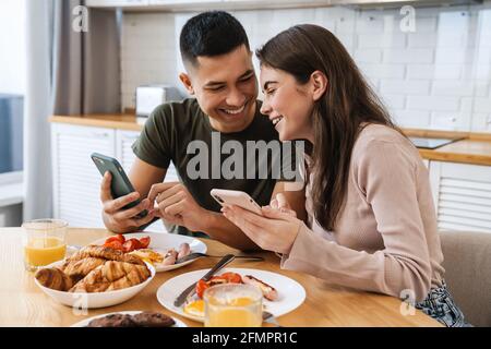 Portrait d'un couple souriant utilisant un smartphone et parlant tout en prenant le petit déjeuner dans une cuisine confortable à la maison Banque D'Images
