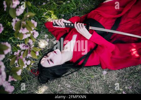 Une jeune femme dépeint la geisha habillée de kimono traditionnel avec une épée de samouraï japonaise dans ses mains et se trouve sur le sol à proximité de sakura en fleurs. Banque D'Images