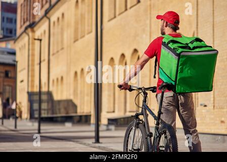 Vue arrière du liveur debout avec vélo et thermo sac sur la rue ensoleillée Banque D'Images