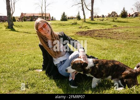 Jeune femme en lunettes de soleil rouges joue avec son corgi sur une pelouse verte d'été. Jour ensoleillé. Un animal de compagnie et un propriétaire heureux Banque D'Images