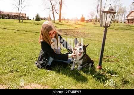 Jeune femme en lunettes de soleil rouges joue avec son corgi sur une pelouse verte d'été. Jour ensoleillé. Un animal de compagnie et un propriétaire heureux Banque D'Images