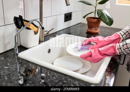 Une femme court rinçant les plats avec une éponge violette au-dessus de l'évier de cuisine, femme de ménage non reconnaissable à la maison faisant des tâches ménagères, portant du caoutchouc Banque D'Images