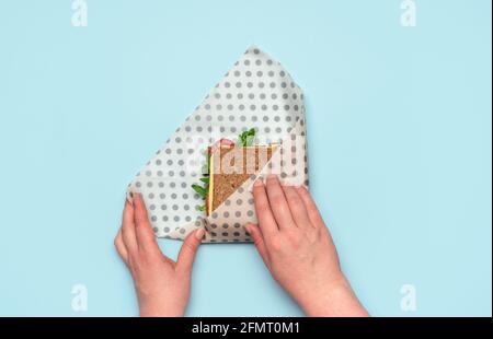 Les mains de la femme enveloppent un sandwich dans un tissu de cire d'abeille sur une table bleue, au-dessus de la vue. Sandwich sain avec du pain complet et des ingrédients végétaliens, enveloppé Banque D'Images