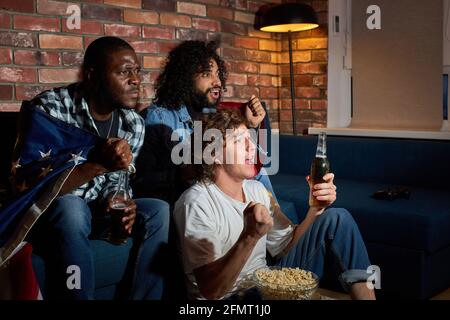 Les gars détendus regardant un match de football à la maison. Groupe multiethnique d'amis, fans applaudissent pour l'équipe nationale préférée, boire de la bière. Concept de Banque D'Images