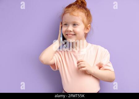 Enfant fille communique sur téléphone mobile, sourit et parle isolé sur fond violet. Gros plan sourire enfant parlant sur smartphone. Les gens sont sincères émoti Banque D'Images