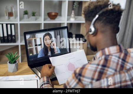 Vue de l'épaule de l'homme africain dans un casque à l'aide d'un ordinateur portable sans fil pour la vidéoconférence avec une femme asiatique. Deux collègues multiraciaux discutent de questions de travail à distance tout en restant à la maison. Banque D'Images