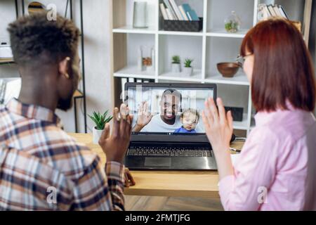 Jeune couple multiculturel ayant un appel vidéo sur un ordinateur portable moderne avec un homme africain qui tient bébé garçon mignon sur les mains. Des amis heureux parlant en ligne tout en restant à la maison.