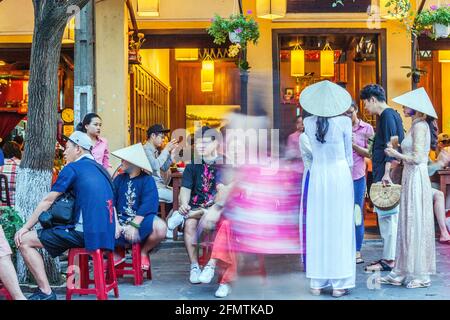 De jolies femmes vietnamiennes portant des chapeaux coniques et des robes traditionnelles d'ao dai se tiennent à l'extérieur du restaurant dans la vieille ville, Hoi an, Vietnam Banque D'Images