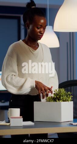 Contrarié congédié une employée de femme noire mettant des choses dans une boîte de la table au bureau comme étant congédiées. Sans emploi emballer des choses tard dans la nuit. Femme fermant l'ordinateur portable et quittant le bureau de travail à minuit Banque D'Images