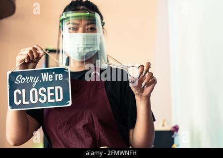 Coiffeur asiatique mignon avec masque oculaire, masque de protection en t-shirt noir et tablier avec une plaque métallique DÉSOLÉ QUE NOUS SOMMES FERMÉS pour à mesure de verrouillage. Banque D'Images