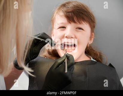 Sourire le visage d'une petite fille sur rendez-vous chez le dentiste. Femme dentiste portant des gants noirs pour vérifier les dents de lait avec miroir et outil explorer. Concept de la dentisterie pédiatrique et des soins dentaires. Banque D'Images