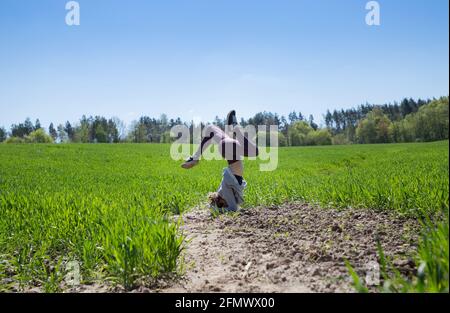 une jeune femme mince se tient à l'envers sur sa tête, tenant son équilibre, parmi l'herbe verte brillante. Mode de vie actif. Exercice en plein air avec santé Banque D'Images