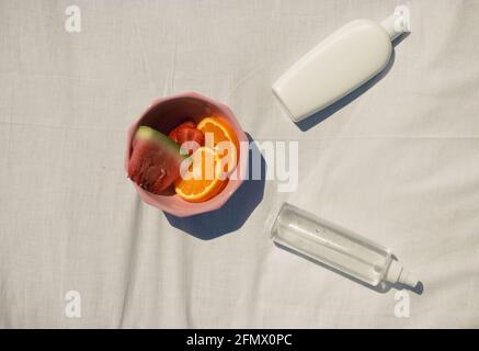 Image horizontale d'un assortiment de fruits dans un bol rose avec une crème solaire et de l'eau hydratante sur un fond de tissu blanc. Concept d'été 2021. Banque D'Images