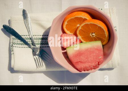 Détail de fruits assortis (oranges, fraises et pastèques) dans un bol rose avec fourchettes et serviette sur fond de tissu blanc. Été 2021 Banque D'Images
