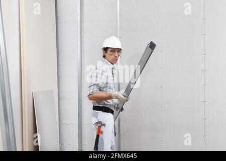 homme travailleur avec des profilés en métal pour cloisons sèches pour l'installation de plaques de plâtre sur le mur. Porter un casque de sécurité blanc, des gants de travail et des lunettes de sécurité. Isolé sur W Banque D'Images