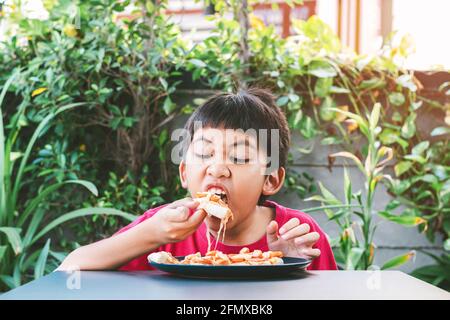 Petit garçon asiatique dans une chemise rouge heureux de manger de la pizza. Banque D'Images