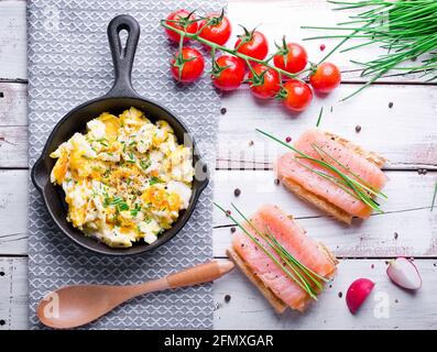 oeufs brouillés avec ciboulette dans une casserole en fonte, toasts avec saumon et ciboulette, et tomates. Des petits déjeuners sains sur une table blanche en bois Banque D'Images