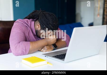 Épuisé Africain-américain homme avec des serrures cheveux prend une sieste sur le lieu de travail, fatigué indépendant gars dort sur la table devant l'ordinateur portable. Concept de surcharge et de surcharge Banque D'Images