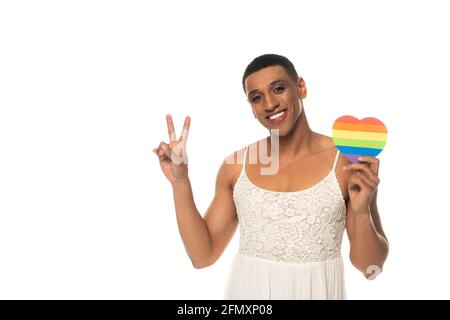 souriant afro-américain transvestite homme tenant lgbt couleurs papier coeur et montrant le signe de la paix isolé sur blanc Banque D'Images