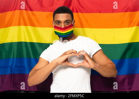 homme afro-américain portant des couleurs arc-en-ciel masque médical et montrant signe du cœur sur le fond du drapeau lgbt Banque D'Images
