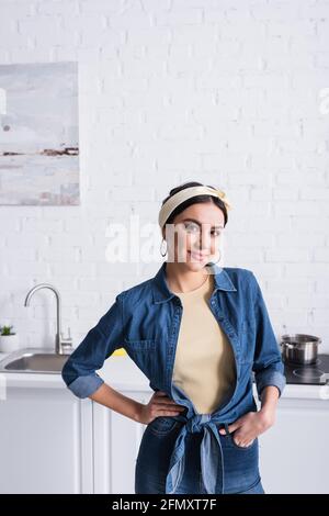 Femme de ménage avec la main sur la hanche souriant à l'appareil photo dans la cuisine Banque D'Images