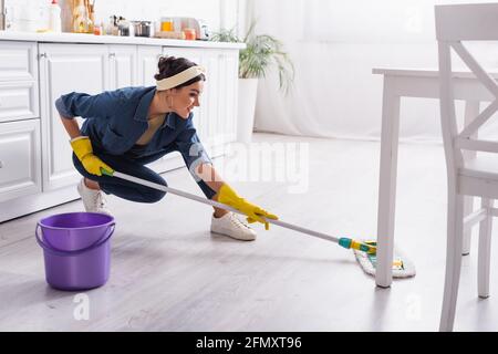 Femme de ménage positive en denim, chemise de lavage, sol de la cuisine Banque D'Images