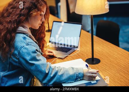Une étudiante qui écrit dans son carnet alors qu'elle étudie à la bibliothèque. Une jeune fille élève prend des notes pour l'examen à la bibliothèque le soir. Banque D'Images