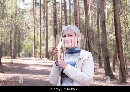 Portrait d'une femme caucasienne âgée en vêtements décontractés mangeant une banane et regardant l'appareil photo sur le fond d'un parc forestier de printemps. Banque D'Images