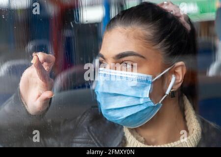Une femme portant un masque de protection face à une fenêtre de bus imbibée de pluie. Banque D'Images