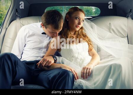 De beaux jeunes mariés qui se embrasent et sourient dans la voiture. Jolie femme aux cheveux rouges vêtue d'une robe blanche, un homme dans la chemise blanche, un noeud papillon noir. Banque D'Images