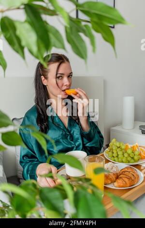 Jeune femme en pyjama de soie a un petit déjeuner sain tout en étant allongé sur le lit dans la chambre. Concept de bien-être à la maison. Santé émotionnelle d'une jeune femme Banque D'Images