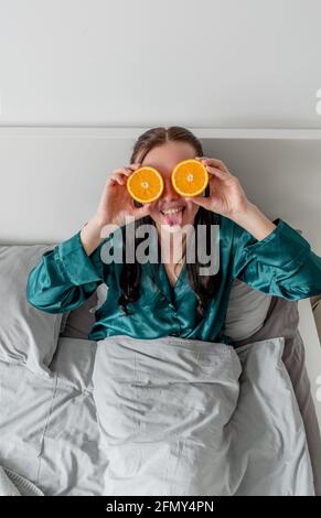 La jeune femme rit et tient les oranges près de ses yeux. Concept de bien-être et zone de confort à la maison. Banque D'Images