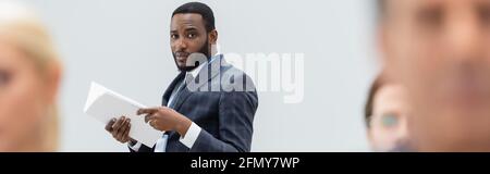 Homme d'affaires afro-américain avec des documents regardant l'appareil photo près de collègues flous, bannière Banque D'Images