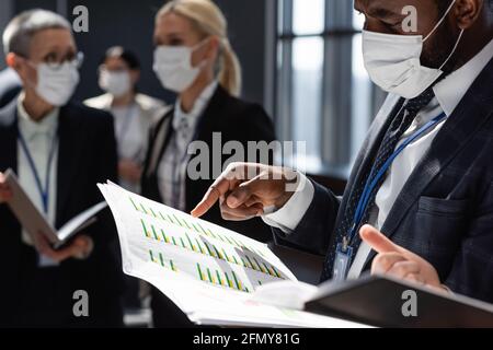 homme d'affaires afro-américain dans un masque médical pointant sur des documents avec graphiques près de collègues flous Banque D'Images