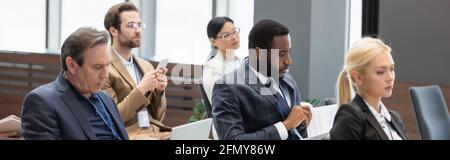 Homme d'affaires afro-américain regardant des journaux près de collègues dans la salle de conférence, bannière Banque D'Images