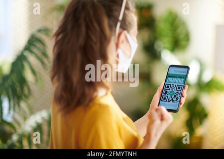 Voyager pendant la pandémie de Covid-19. Vue de derrière une femme voyageur seule avec un smartphone, une application de passeport covid et un masque médical dans le salon au soleil Banque D'Images