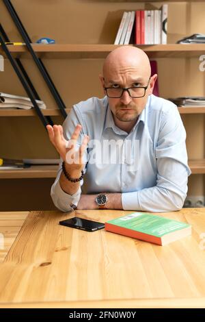 un homme d'âge moyen et chauve avec des lunettes est assis à une table pour poser une question, menant la discussion tout en regardant la caméra Banque D'Images