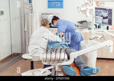 Dentiste senior femme effectuant la chirurgie mettant l'implant dans la bouche de l'homme malade dans la clinique dentaire. Médecin dentaire avec masque de protection traitant les dents du patient assis sur la chaise pendant la consultation Banque D'Images