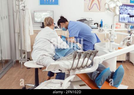 Homme malade assis sur une chaise dentaire pendant un examen médical alors qu'une femme dentiste senior effectue une chirurgie buccale au bureau de la dentisterie. Équipe d'hôpital examinant le mal de dents du patient préparant le traitement dentaire Banque D'Images