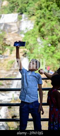 Ella, Sri Lanka - 04 15 2021: Enfant tenant un smartphone au-dessus de sa tête et prendre une photo de selfie devant la chute d'eau de Rawana Ella. Banque D'Images