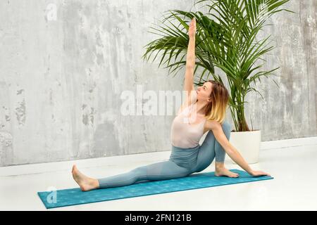 Femme faisant un Upavista Konasana variation yoga pose l'étirant bras au-dessus de sa tête avec un mur gris et en pot toile de fond de plante dans une santé et fitn Banque D'Images