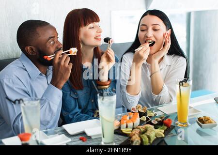 Les gens, la nourriture, le plaisir, le repas de sushi. Gros plan de trois amis multiethniques affamés et excités, s'amuser dans un café, manger de la nourriture asiatique, prendre des petits pains à sushis avec des bâtonnets de bambou Banque D'Images