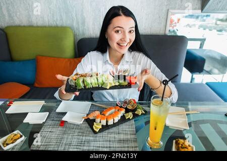 Jeune femme asiatique brune en vêtements décontractés tient dans les mains des petits pains à sushis servis sur une assiette noire, assis dans un restaurant japonais traditionnel. Les gens, le style de vie et le concept de la nourriture asiatique Banque D'Images